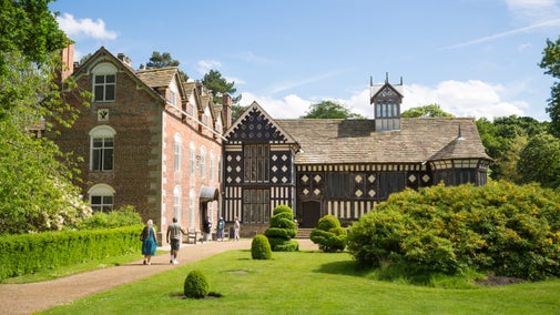 Two visitors are walking past a bright green lawn which has decorative bushes planted on it, towards the house at Rufford Old Hall, Lancashire, on a bright sunny day.